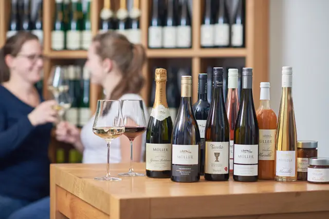 Weinsortiment eines Weinguts auf einem Holztisch in der Vinothek, im Hintergrund zwei Frauen bei der Verkostung