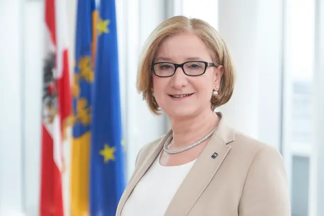 Freundliches Politikerin-Portrait im beigen Blazer vor Fahnen in hellem Büro