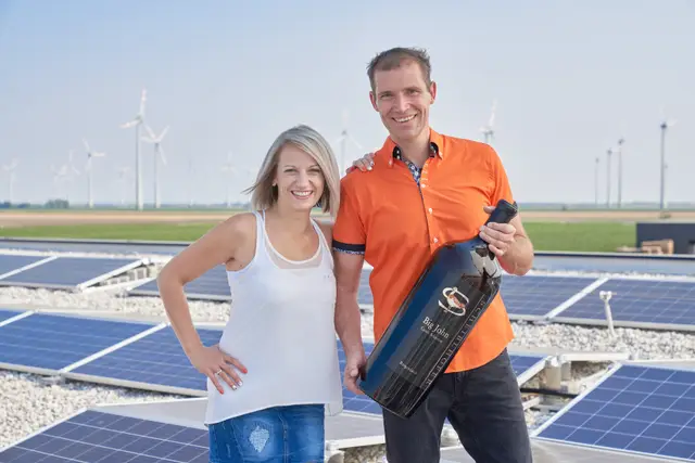 Lachendes Winzerpaar mit großer Weinflasche auf dem Photovoltaikdach ihres Betriebs, dahinter Windräder im Weinviertel