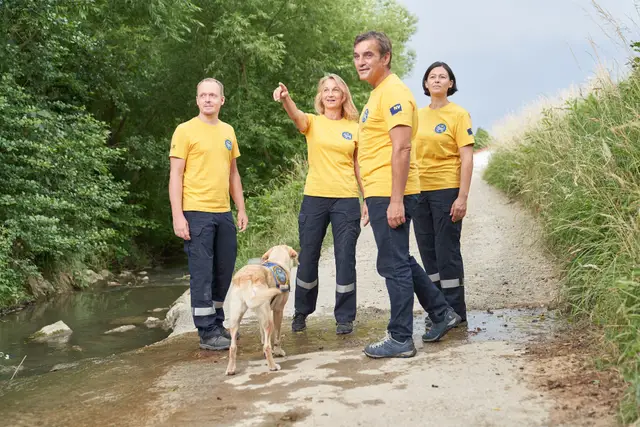 Vierköpfiges Einsatzteam in gelben Shirts mit Suchhund an einem Bachlauf, Teamfoto im Freien