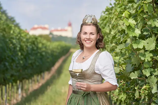 Weinkönigin im Dirndl mit Krone und Weinglas zwischen den Rebzeilen, im Hintergrund das Stift Göttweig