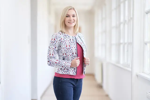 Junge Politikerin im gemusterten Blazer lächelt in hellem Altbaugang – natürliches Kandidatinnen-Portrait