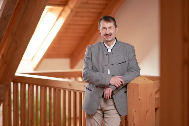 Unternehmer im grauen Trachtensakko lehnt entspannt an einem Holzgeländer im hellen Dachgeschoss