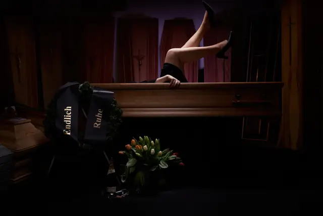 Beine in Pumps ragen aus einem Sarg, davor Trauerkranz und Sekt – Kampagnenmotiv mit schwarzem Humor