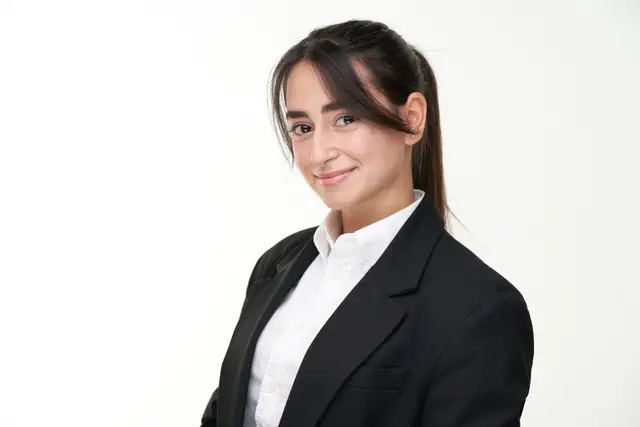Junge Frau mit Pferdeschwanz im schwarzen Blazer und weißer Bluse, sanft lächelnd vor weißem Hintergrund, Bewerbungsfoto im Studio