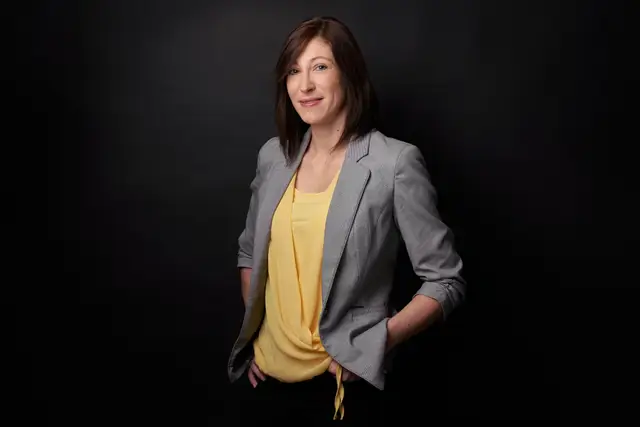 Frau im grauen Blazer mit gelbem Top, Hände lässig in den Taschen, Business-Portrait vor dunklem Studiohintergrund