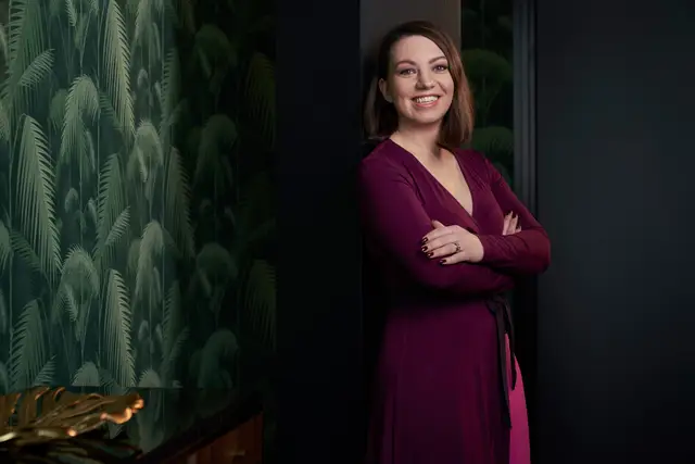 Lächelnde Unternehmerin im weinroten Wickelkleid mit verschränkten Armen vor dunkler Wand mit Palmenmotiv