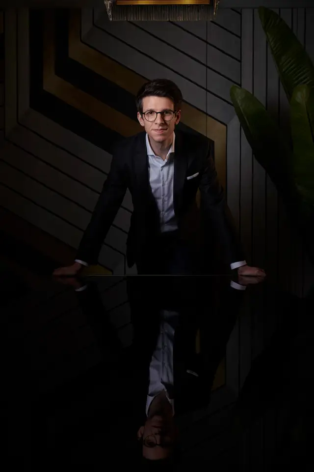 Junger Unternehmer mit Brille stützt sich auf einen spiegelnden Tisch, stilvolles dunkles Interieur
