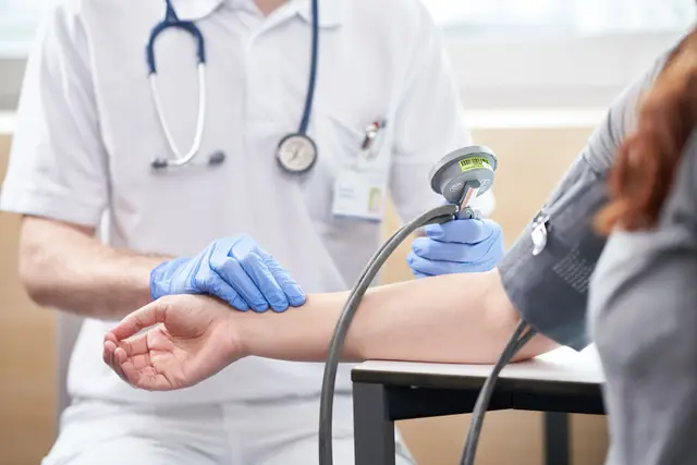 Blutdruckmessung in der Ordination, Nahaufnahme der behandschuhten Hände am Arm der Patientin