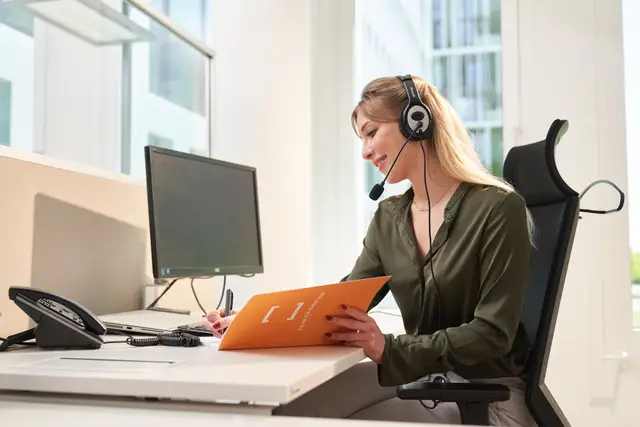 Mitarbeiterin mit Headset macht lächelnd Notizen am Schreibtisch im hellen Büro – Imagefoto am Arbeitsplatz
