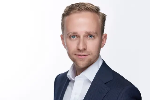 Mann mit feiner Brille in dunkelblauer medizinischer Berufskleidung, freundlicher Blick vor blauem Studiohintergrund