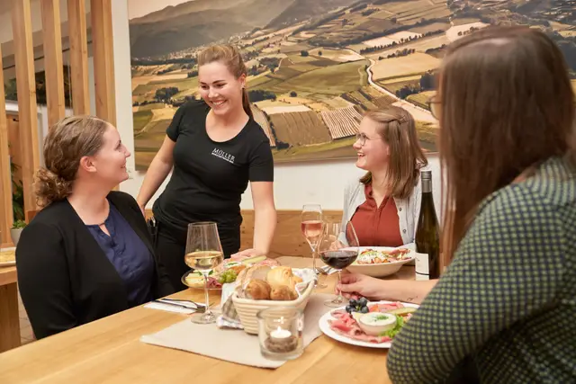 Servicemitarbeiterin serviert lächelnd Wein und Brettljause an einen Tisch mit Gästen in der Vinothek