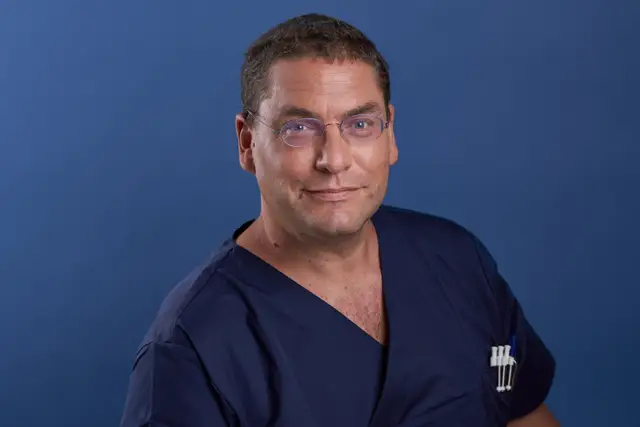 Herzlich lachender Mann mit markanter Brille im grauen Pullover vor weißem Hintergrund, modernes Business-Portrait