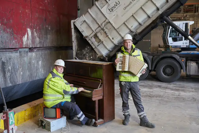 Zwei Mitarbeiter in Warnwesten musizieren mit Klavier und Akkordeon in einer Recyclinghalle, humorvoll inszenierte Aufnahme
