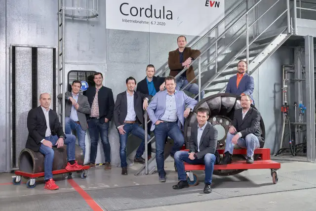 Zehnköpfiges Team locker gruppiert in einer Kraftwerkshalle mit Stiege und Turbinenrad – Teamfoto in der Produktion