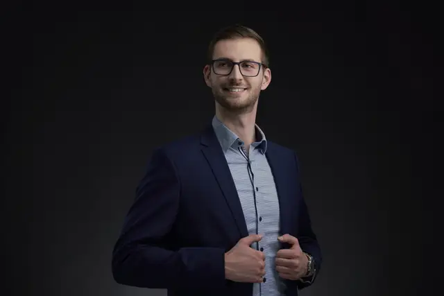 Lächelnder junger Mann mit Brille im dunkelblauen Anzug und hellblauen Hemd vor schwarzem Hintergrund, Business-Portrait