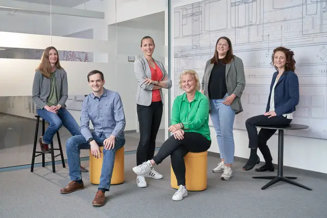 Sechs Mitarbeitende entspannt gruppiert im modernen Büro vor einer Wand mit technischen Zeichnungen