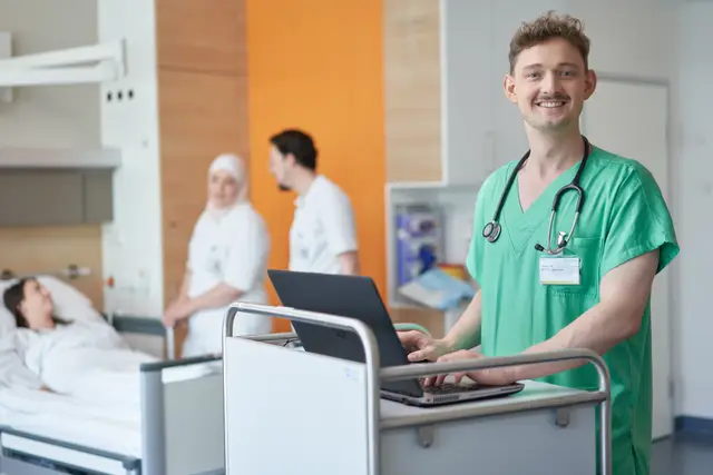 Lächelnder junger Mediziner in grünen Scrubs mit Stethoskop am Visitenwagen im Patientenzimmer