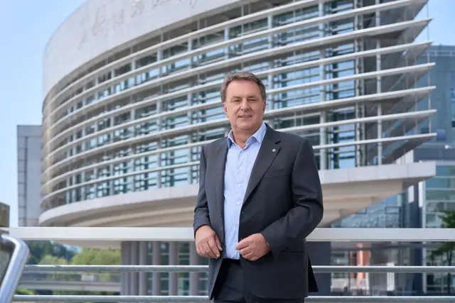 Politiker im grauen Sakko lehnt an Geländer vor moderner Glasfassade – souveränes Portrait im Regierungsviertel