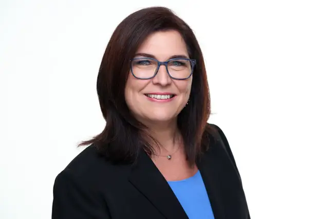 Frau mit blauer Brille im schwarzen Blazer und blauem Top, warm lächelnd vor weißem Hintergrund, Bewerbungsfoto im Studio