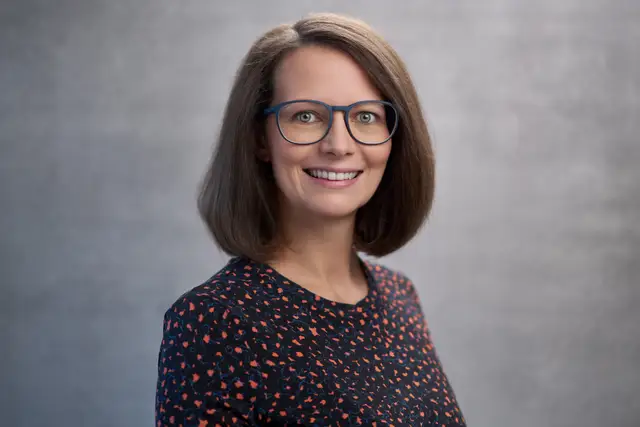 Lächelnde Frau mit Brille und braunem Bob, Mitarbeiterportrait vor grauem Studiohintergrund