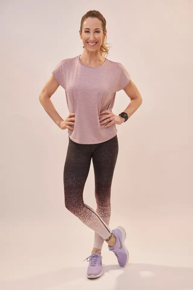 Sportliche Frau in Trainingsoutfit mit Leggings und Laufschuhen, lachend in Ganzkörperpose vor rosé getöntem Studiohintergrund