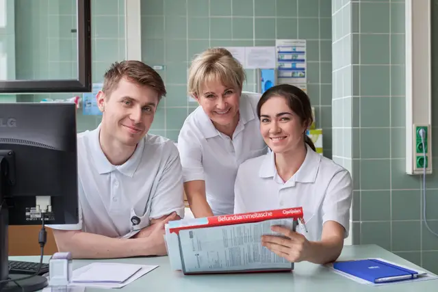 Teamfoto von drei Pflegekräften mit Befundmappe am Stützpunkt der Station