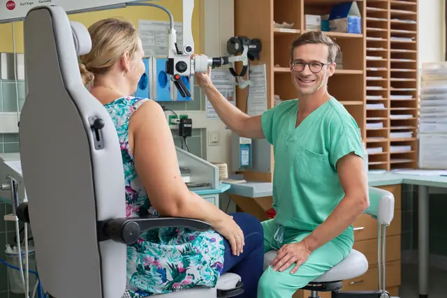 HNO-Arzt in grünen Scrubs am Ohrmikroskop, lächelnd neben seiner Patientin in der Ambulanz