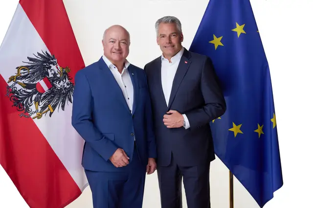Zwei Politiker in dunkelblauen Anzügen posieren lächelnd zwischen österreichischer und europäischer Fahne