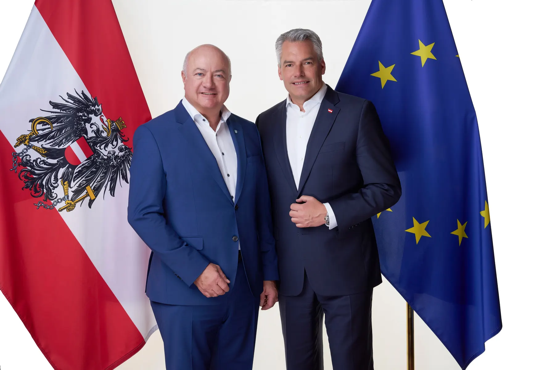 Zwei Politiker in dunkelblauen Anzügen posieren lächelnd zwischen österreichischer und europäischer Fahne