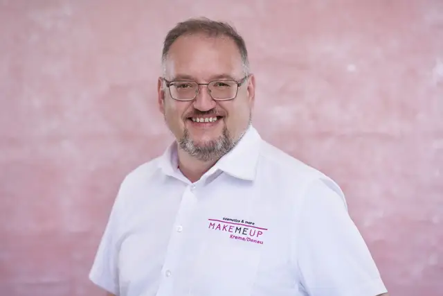 Herzlich lachender Mann mit Brille und grauem Bart im weißen kurzärmeligen Hemd vor rosa strukturiertem Studiohintergrund