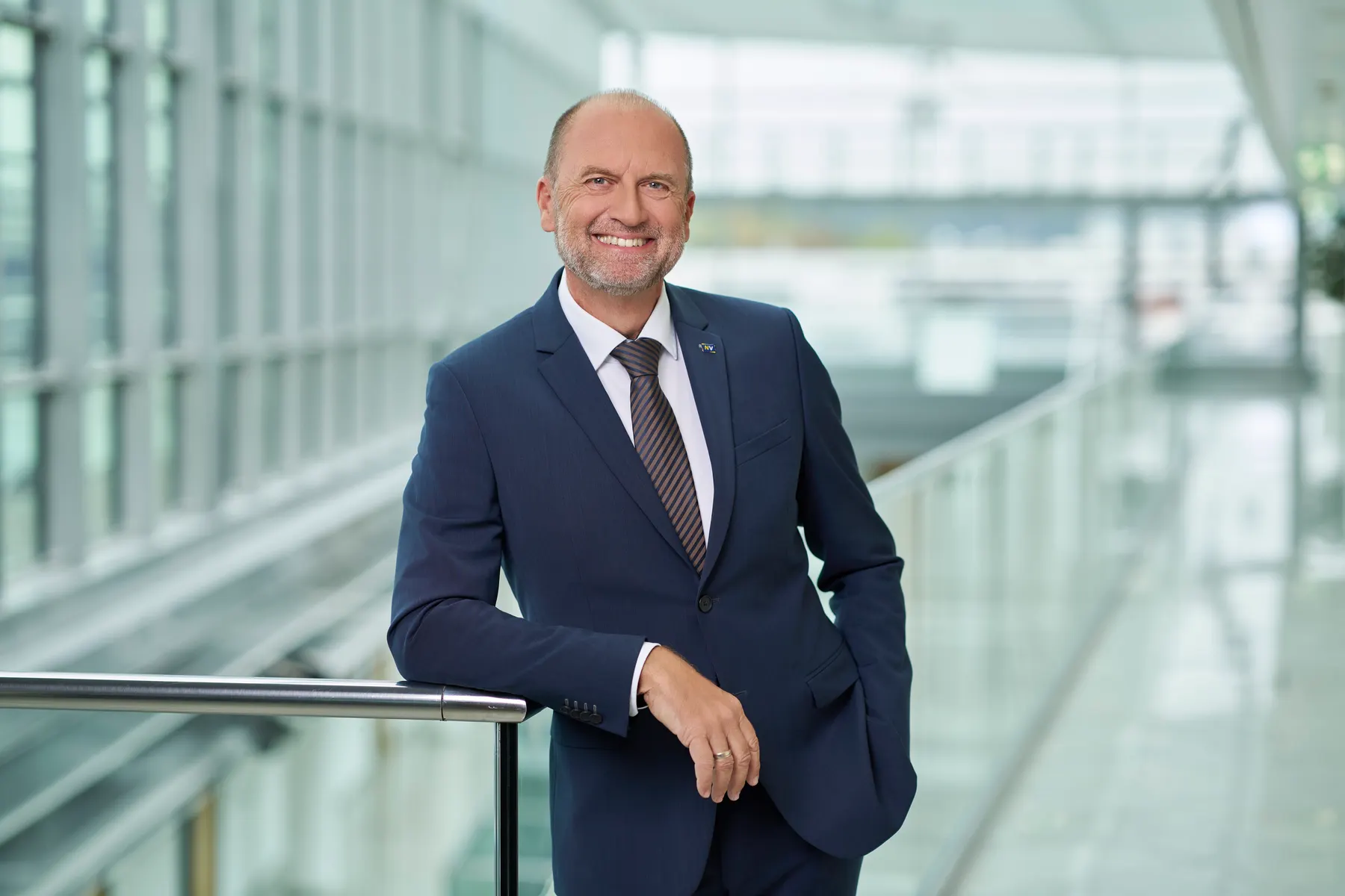 Strahlender Geschäftsführer im dunkelblauen Anzug, an ein Geländer gelehnt, in lichtdurchflutetem Bürogebäude