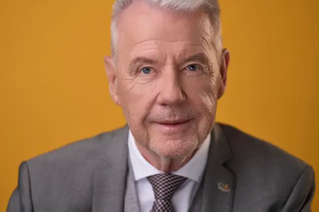 Nahes Portrait eines grauhaarigen Politikers im grauen Anzug vor gelbem Hintergrund – markantes Plakat-Sujet