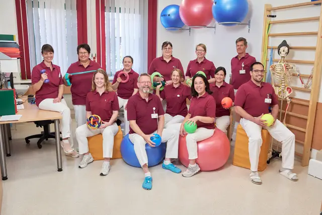Physiotherapie-Team in weinroten Polos, Teamfoto mit Gymnastikbällen im Therapieraum