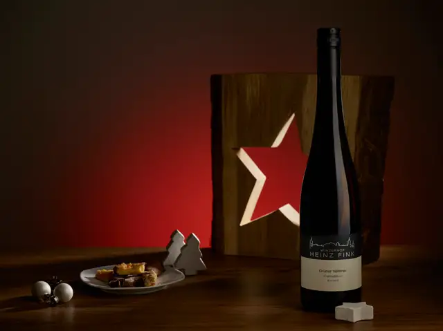 Weinfotografie im Advent: Flasche Grüner Veltliner vor beleuchtetem Holzstern mit Keksen und Christbaumkugeln
