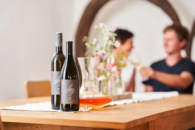 Zwei Weinflaschen mit Dekanter auf dem Verkostungstisch, Gäste bei der Weinprobe im Hintergrund