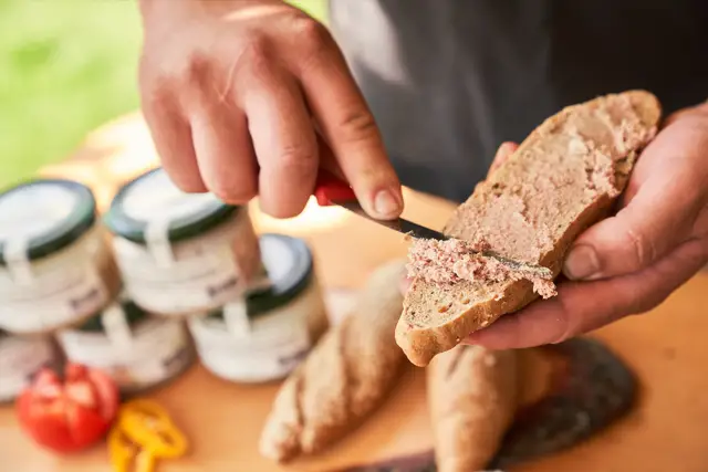 Hände streichen hausgemachten Aufstrich aufs Brot, Gläser der Hofmanufaktur im Hintergrund