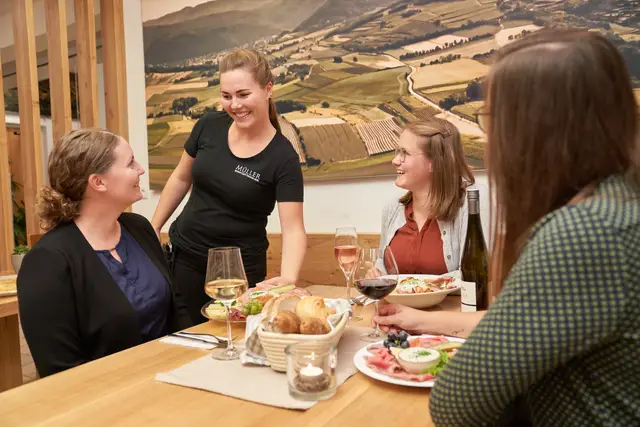 Gastgeberin serviert lachenden Gästen Jausenplatten und Wein, herzliche Stimmung im Buschenschank