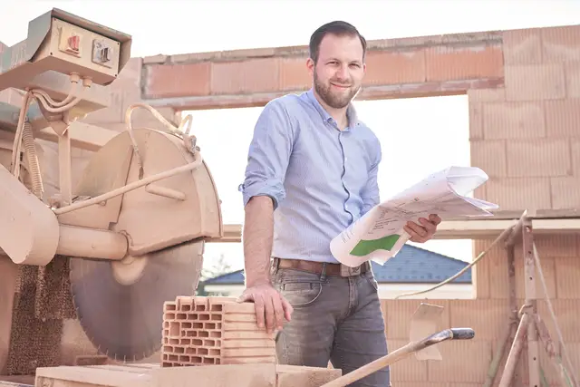 Junger Baumeister mit Bauplänen im Rohbau, hinter ihm die Steinsäge – Reportage von der Baustelle