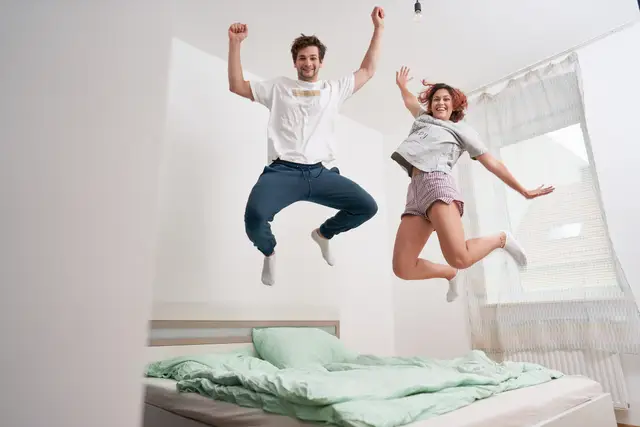 Junges Paar springt jubelnd auf dem Bett der neuen Wohnung – Lebensfreude pur