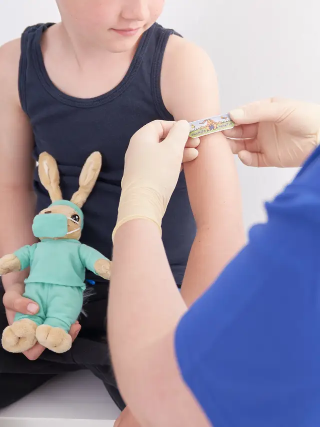 Ein Bub mit Stoffhase bekommt in der Ordination ein buntes Pflaster auf den Arm geklebt