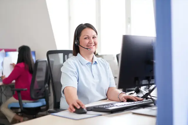 Mitarbeiterin mit Headset am Schreibtisch im Kundenservice, freundlicher Blick in die Kamera