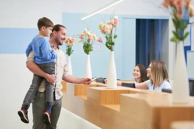 Vater mit Kind auf dem Arm beim freundlichen Empfang an der Rezeption einer Klinik