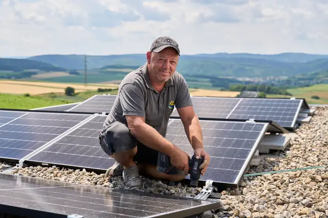 Monteur verschraubt Photovoltaik-Paneele am Flachdach, dahinter das hügelige Umland – Reportage