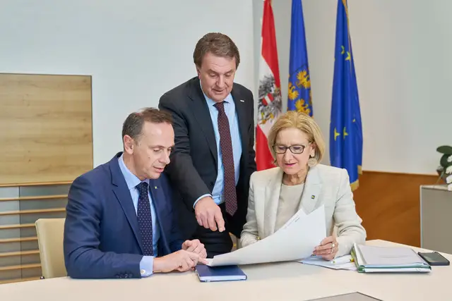 Drei Regierungsvertreter besprechen Unterlagen vor Österreich-, Landes- und EU-Fahne im Amtsgebäude