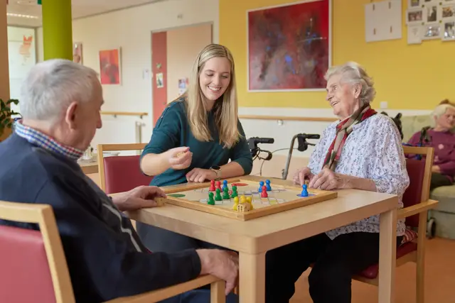 Betreuerin spielt mit Bewohnern im Pflegeheim eine Runde Brettspiel, herzliche Stimmung am Tisch