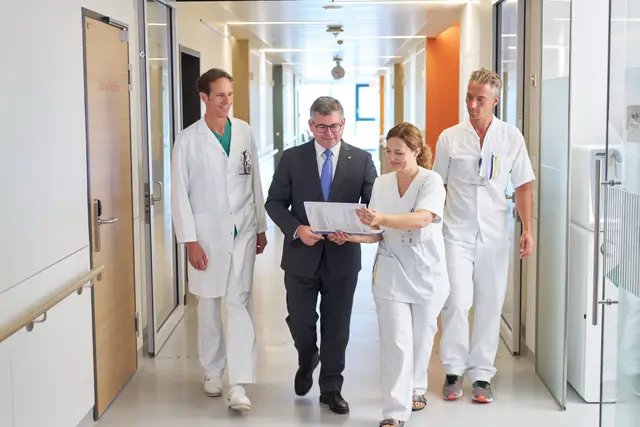 Führungskraft im Anzug bespricht mit Ärzten und Pflegekräften Unterlagen am Gang des Krankenhauses