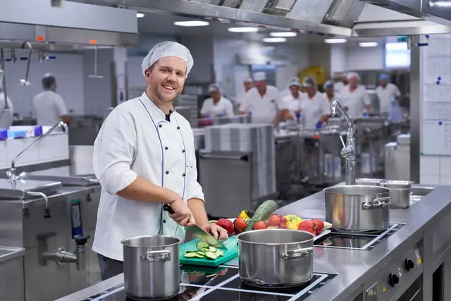 Koch schneidet frisches Gemüse in der Großküche, dahinter das Küchenteam bei der Arbeit