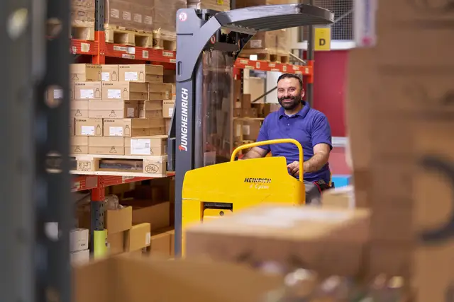 Lagermitarbeiter am Hochhubwagen zwischen Palettenregalen, Reportage aus der Logistik