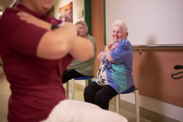 Seniorinnen turnen mit der Therapeutin im Sitzen, gelöste Stimmung bei der Gymnastik im Pflegeheim